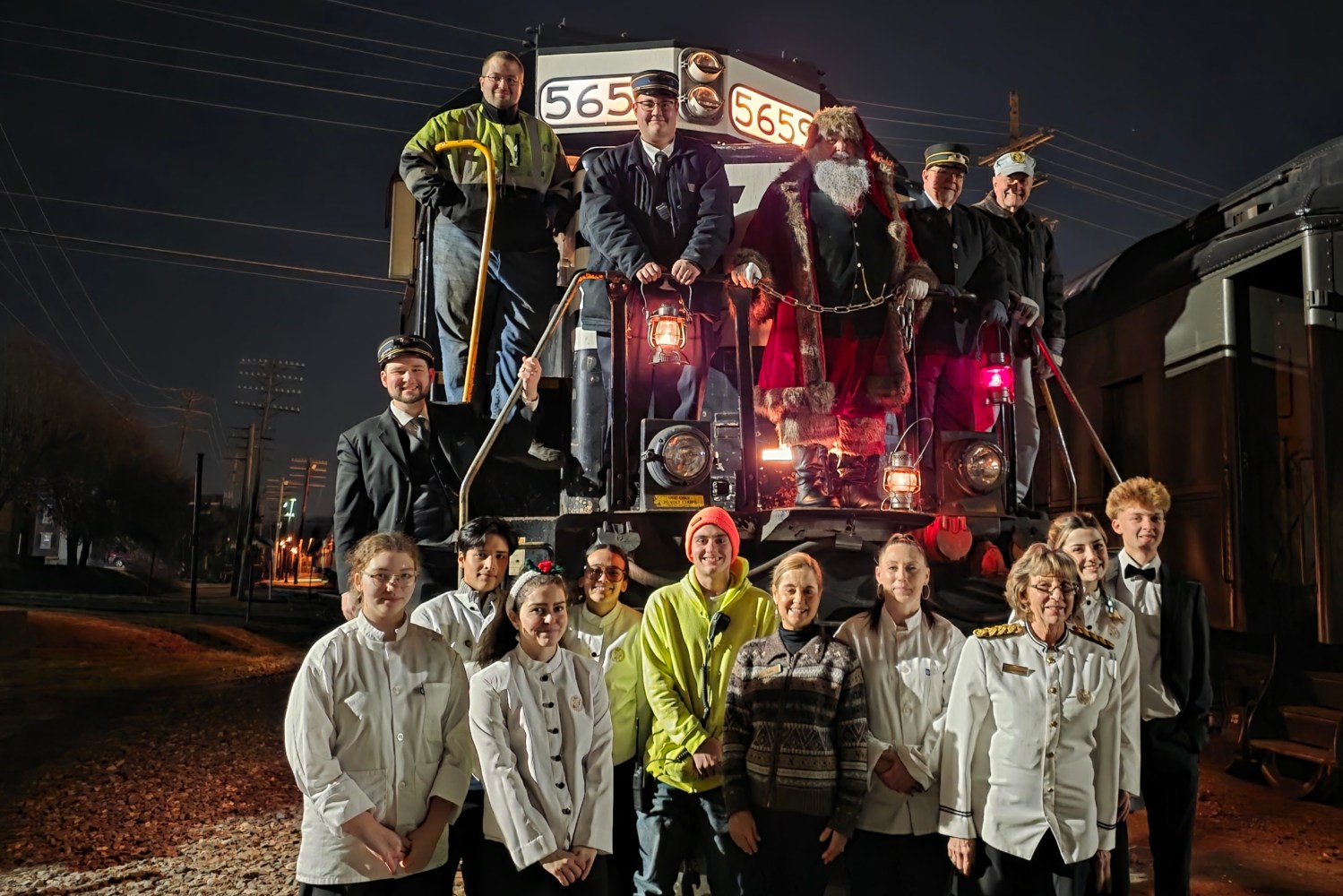 Group of people posing in front of a train at night, including a person dressed as Santa Claus.