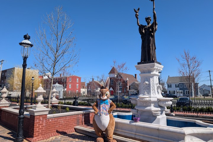 Mascot in bunny costume by statue in park with blue sky background.