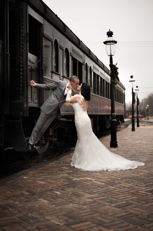 Bride and groom share a kiss beside a vintage train on a cobblestone platform.